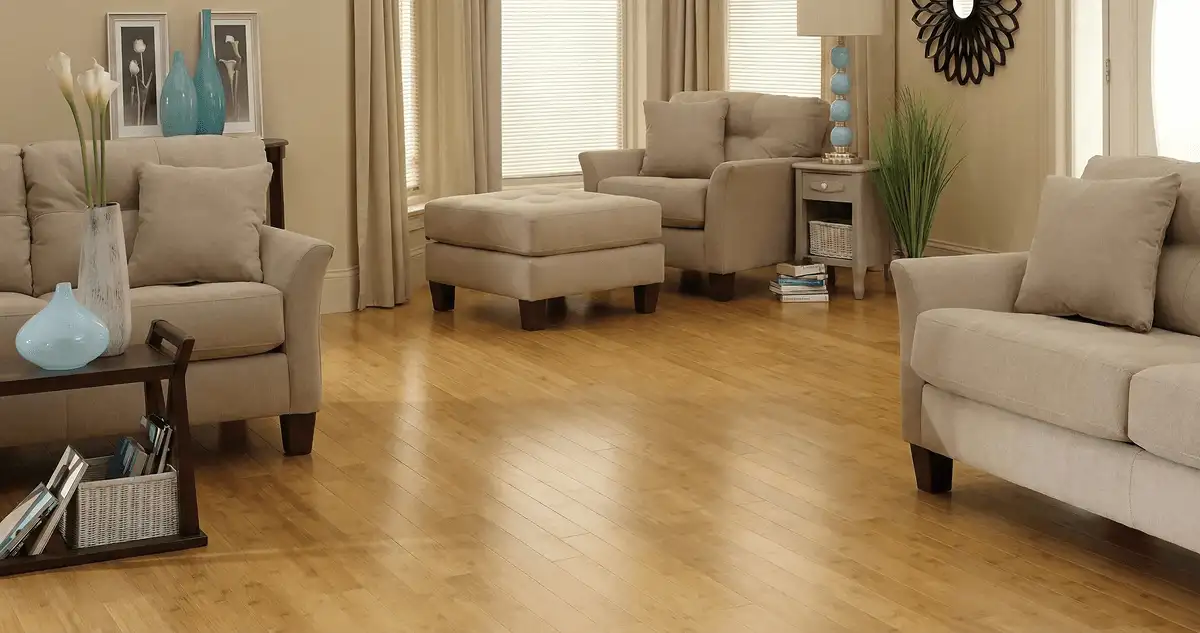 A living room with professionally installed bamboo flooring, beige sofas, a matching ottoman, a side table with a blue lamp, vases, framed pictures, and large windows with blinds and curtains in Huntsville, AL